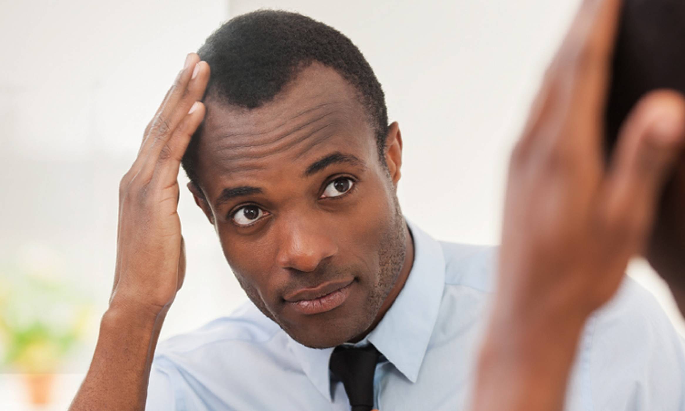 A handsome black man looks at his reflection in the mirror as he touches his scalp. He has short hair with a receding hairline.