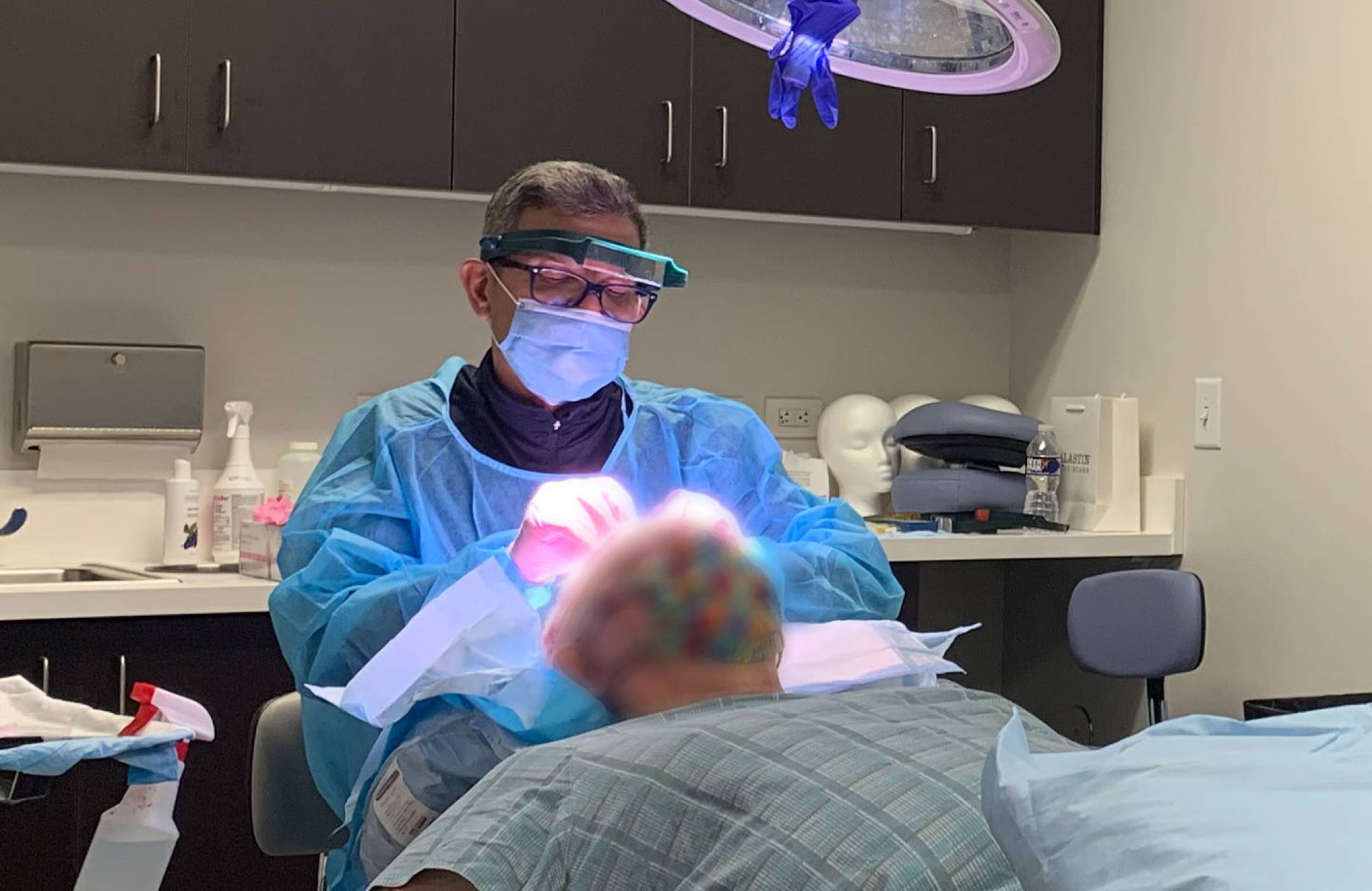 Dr. Zack Charkawi is performing a hair transplant surgery in the operating room of the clinic. Two of his team
                            remembers are working by his side. The patient's face is blurred out.