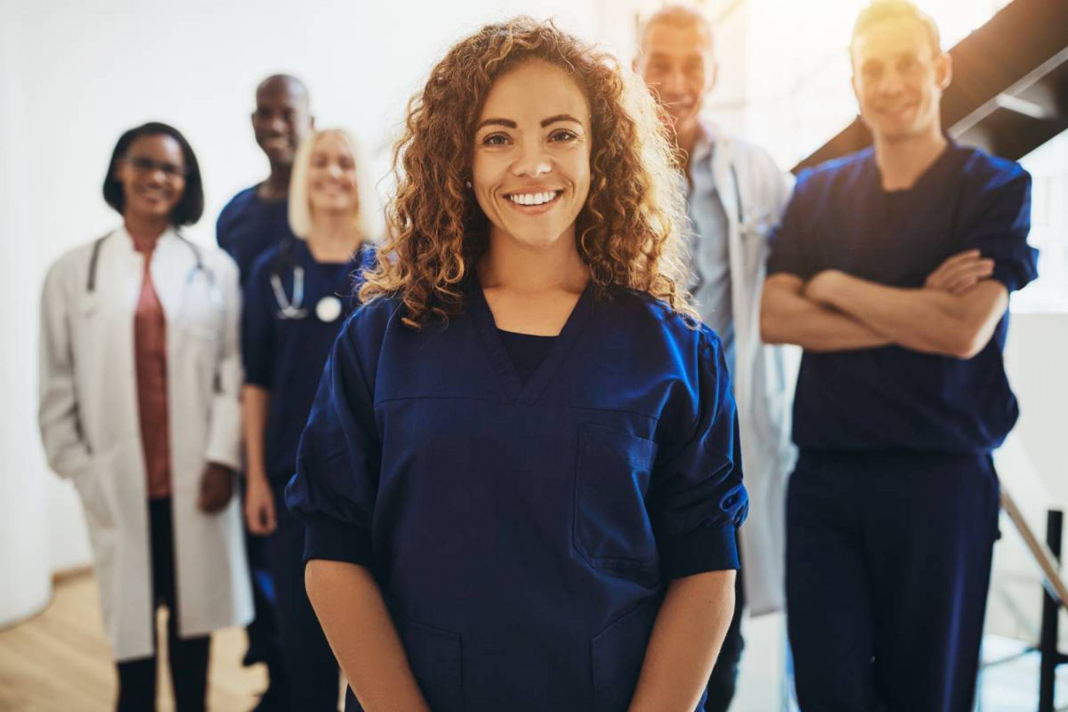 A standing female nurse is looking directly at us with a warm smile. She is surrounded in the back by a medical team of male and female doctors and nurses.